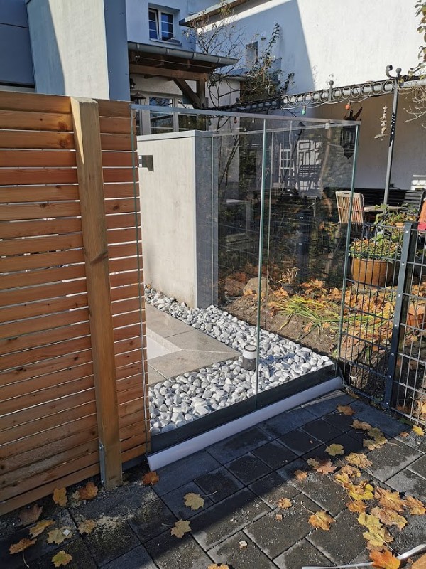 Eine Gartenecke, statt eines Zaunes wurden große Glasscheiben verwendet, sodass man auf ein Steinbeet schaut.
