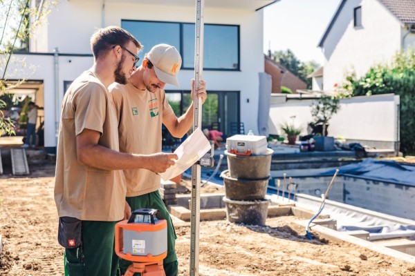Zwei Arbeiter, die sich einen Plan anschauen. Sie stehen in einer Gartenbaustelle.