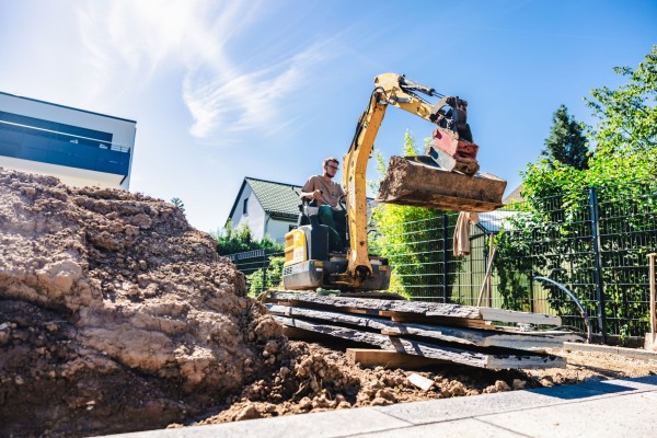 Ein Mann fährt in einem Bagger über eine Gartenbaustelle, links ist ein großer Erdhaufen.