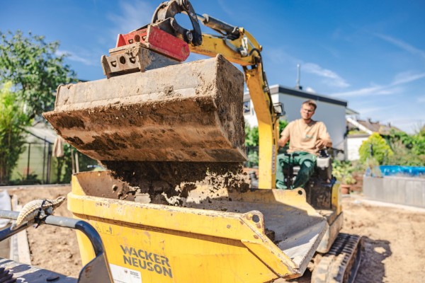 Ein kleiner gelber Gartenbagger, der von einem jungen Mann gesteuert wird, lässt über die Schaufel Dreck in einen gelben Container fallen.