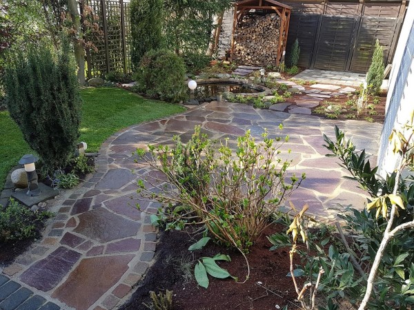 Ein garten mit einer Terasse aus unterschiedlich großen rötlichen Natursteinen.