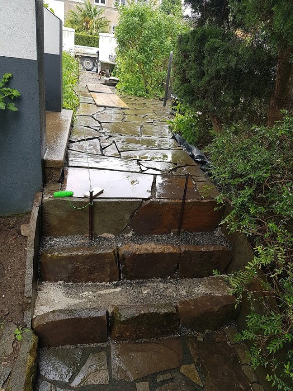 Eine Treppe aus großen Natursteinen in unterschiedlichen Größen, die Steine sind nass vom Regen. Der Weg oben ist aus den gleichen Steinen gepflastert.