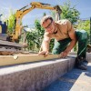 Ein junger Mann kniet auf Beton und überprüft mit einer Wasserwaage die gelegten grauen Steine. Er lächelt und schaut in die Kamera. Im Hintergrund ist ein gelber Bagger zu sehen.