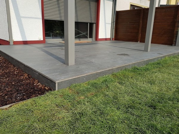 Eine Terasse aus grauen Steinplatten, davor eine Rasenfläche und links daneben ein Abschnitt mit Rindenmulch.