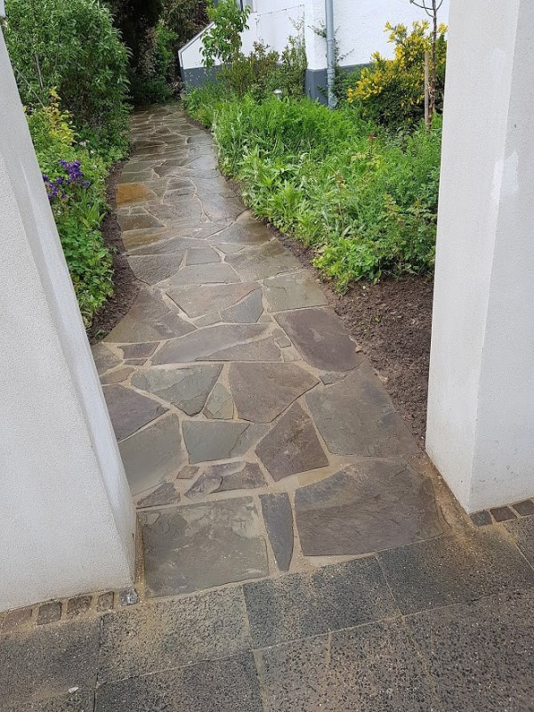 Ein Weg in einem Garten, aus verschiedengroßen Natursteinen gepflastert. Links und rechts davon sind Beete mit Grünpflanzen.