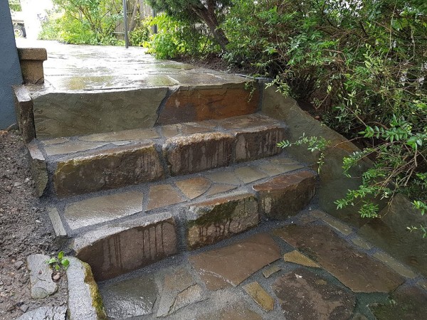 Eine Treppe aus großen Natursteinen in unterschiedlichen Größen, die Steine sind nass vom Regen. Der Weg oben ist aus den gleichen Steinen gepflastert.