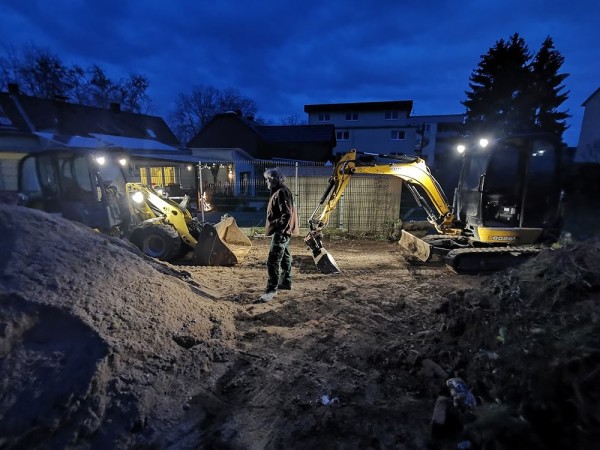 Zwei Bagger, die Nachts einen Garten umgraben. Man sieht große Erdberge. Ein Mann steht in der Mitte des Bildes.