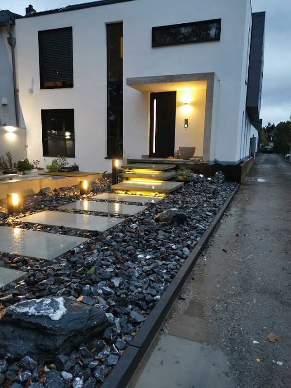 Ein Hauseingang zu dem große, fast schwebende Steinplatten als moderner Treppenersatz führen. Die vorderen Platten sind eingefasst von einem Steinbeet und moderner Gartenbeleuchtung.