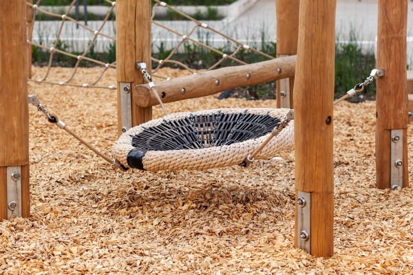 Eine Rundschaukel in der Mitte von vier Holzpfeilern, der Untergrund ist sandfarbener Mulch.
