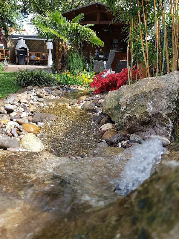 Ein natürlich aussehender kleiner Fluss in einm Garten. Im Flussbett sind kleine Steine, am Rand große. Dahinter sieht man Gartenhaus und Haus.