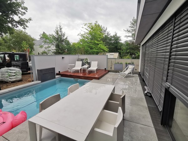 Ein moderner Garten ohne Rasenfläche. Die Hälfte sind graue Steinplatten mit einer Tischgarnitur mit 4 Stühlen, die andere Hälfte sind ein Pool mit einem Holzdeck auf dem zwei Liegen stehen. Im Pool ist ein pinker Flamingo-Schwimmring.