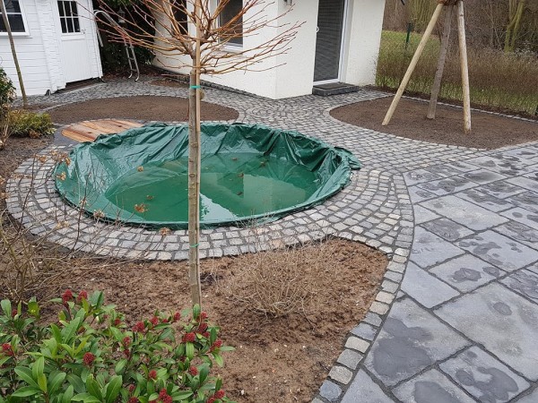 Ein kleiner Garten mit einem, mit einer mit Wasser vollgelaufenen grünen Plane abgedeckten Pool, umringt von Pflastersteinen und Beeten.