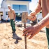 Eine Gartenbaustelle. Nahaufnahme von Händen, die mit einem Hammer einen Pfeiler in den Boden schlagen, im unscharfen Hintergrund sieht man einen weiteren Mitarbeiter der einen Pfeiler bereithält.