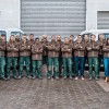 Das Team steht in Arbeitskleidung mit verschränkten Armen und lächelt in die Kamera. Sie stehen vor großen Garagentoren. Hinter dem Team stehen zwei Autos und auch neben dem Team stehen rechts und links jeweils ein Auto.